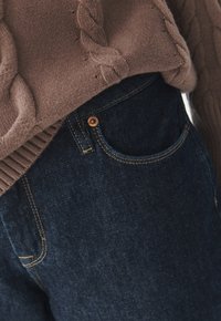 Suéter de punto marrón con un patrón de trenzas, combinado con jeans de mezclilla azul oscuro que presentan un bolsillo frontal y detalles de botones de latón.