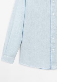Chemise bleu clair à manches longues en tissu tissé. Dotée d'un col, de manches longues et de cinq boutons blancs le long de la patte de boutonnage.