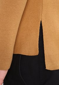 Musztardowy, dzianinowy sweter o fakturowanej powierzchni i bocznym rozcięciu, noszony na czarnych spodniach. Projekt charakteryzuje się prostym krojem i prostymi wykończeniami.