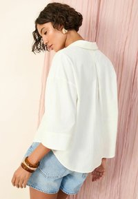 Blusa bianca a maniche corte con colletto, vestibilità rilassata, dettaglio della piega posteriore, abbinata a pantaloni corti in denim e braccialetti trasparenti. Lo sfondo è di un rosa tenue.