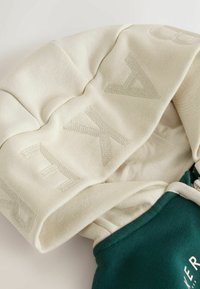 Sudadera con capucha que presenta una capucha beige con letras texturizadas y un cuerpo verde con un logo impreso. Tela suave, ajuste relajado.