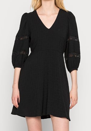 Robe noire avec un décolleté en V et des manches bouffantes. Le tissu présente un motif texturé et des détails en dentelle sur les manches. Ourlet évasé.
