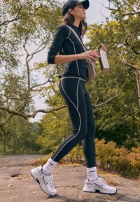 Melna sporta augšdaļa ar baltu šuvju dizainu, apvienota ar melniem pieguļošiem legingiem, baltiem Nike sporta apaviem un potīšu garuma zeķēm, tur roku dzelzs tērauda ūdens pudeli.