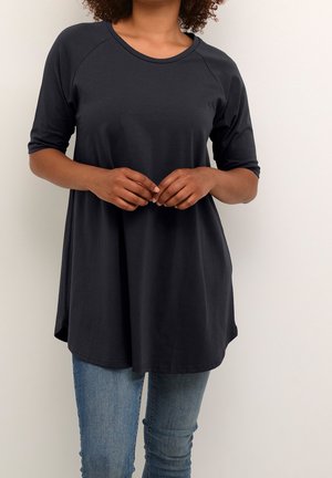 Femme portant un haut noir ample à manches trois-quarts et un jean bleu, debout avec les doigts doucement croisés devant elle.