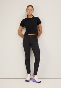 Svart kortärmad t-shirt kombinerad med åtsittande svarta byxor med dragkedjsfickor och logotyp, kompletterat med vita och lila sportskor.