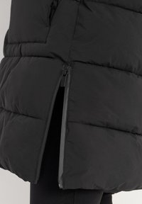 Zwarte gewatteerde jas met een gedeeltelijk open zijrits, waardoor donkere broek eronder zichtbaar is.