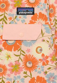 Produto Patagonia com um design floral em laranja, rosa e azul sobre um fundo rosa claro, com um bolso de botão em rosa pálido e uma etiqueta de logotipo.