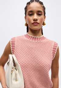 Νεαρή γυναίκα με κοτσίδες, που φοράει ροζ υφασμάτινο πλεκτό τοπ χωρίς μανίκια και χρυσά σκουλαρίκια, κρατώντας μια λευκή τσάντα στον ώμο της.