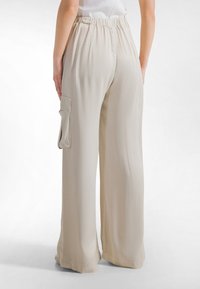 Beige wijde broekspijpen gemaakt van gladde stof; voorzien van een elastische tailleband en een zijzak met klep voor extra functionaliteit.