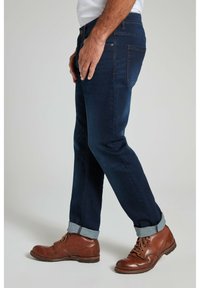 Jeans oscuros de corte slim, con puños enrollados y costuras en contraste, combinados con botas de cuero marrón y una camisa de color claro.