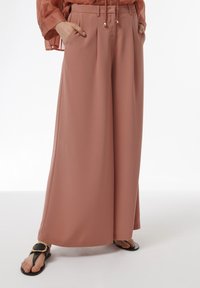 Brede broek in een zachte rozenkleur, gemaakt van vloeiende stof. Kenmerkend door plooien en zakken, gecombineerd met zwarte sandalen en een doorzichtige blouse.