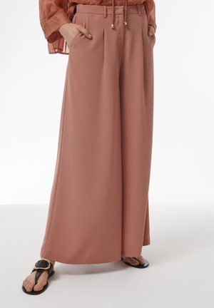 Weite Hosen in einem sanften Rosaton, aus fließendem Material gefertigt. Mit Falten und Taschen, kombiniert mit schwarzen Sandalen und einer transparenten Bluse.