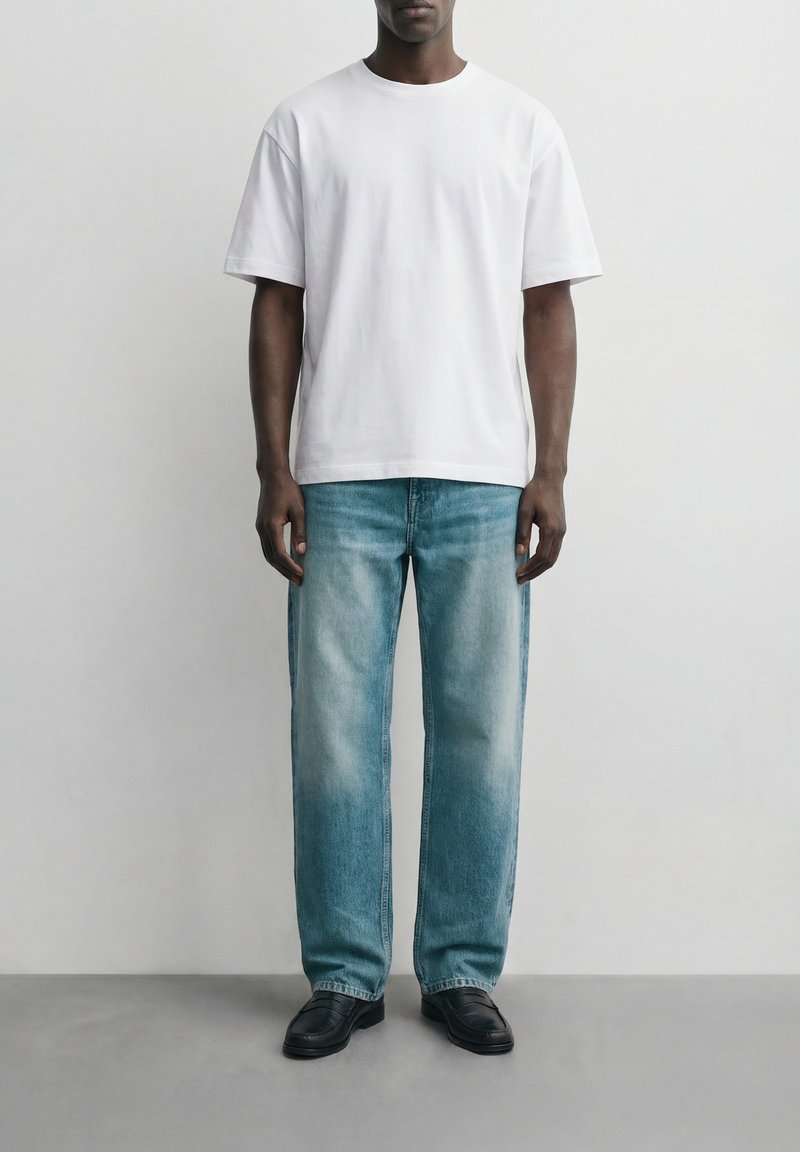 Άνδρας που στέκεται φορώντας απλό λευκό κοντομάνικο μπλουζάκι, ανοιχτό μπλε τζιν με ίσια μπάτζα και μαύρα δερμάτινα παπούτσια, μπροστά σε απλό λευκό φόντο.