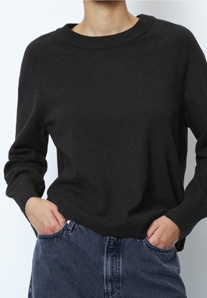 Osoba nosząca czarny sweter z długim rękawem i okrągłym dekoltem oraz niebieskie dżinsy, ręce włożone w przednie kieszenie.