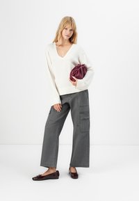 Nainen, joka käyttää valkoista V-kaula-aukkoista neuletta ja harmaita leveälahkeisia cargo-housuja, pitää kädessään taiteltua burgundinpunertavaa clutch-laukkua, seisoo valkoista taustaa vasten.
