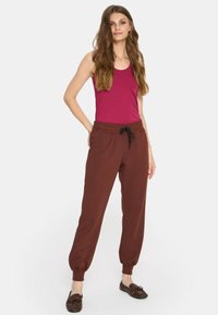 Młoda kobieta z długimi faluj ącymi włosami, ubrana w bezrękawnik w kolorze magenty, brązowe spodnie dresowe z troczkiem i brązowe mokasyny, stoi w zrelaksowanej pozycji.