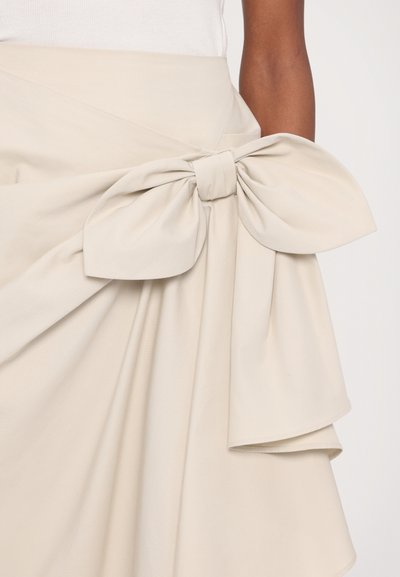 Jupe beige clair avec un grand nœud à la taille, au design structuré et drapé, en tissu lisse et légèrement texturé.