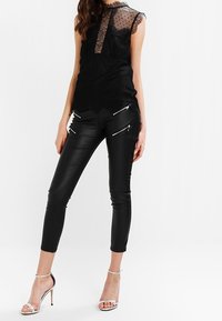Schwarzes Spitzentop mit transparentem Hals und kurzen Ärmeln, kombiniert mit schwarzen Kunstlederhosen mit silbernen Reißverschlüssen und einer schlanken Passform.