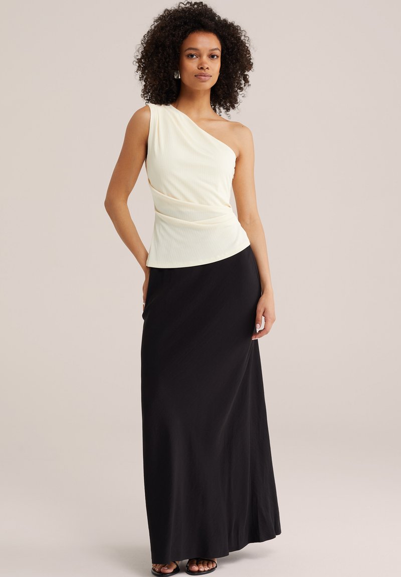 Femme aux cheveux bouclés portant un haut blanc asymétrique sans manches et une longue jupe noire, debout avec une main sur la hanche.