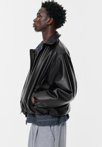 Črna usnjena jakna z urejenim dizajnom, visokim ovratnikom in elastičnimi manšeti. Nosijo jo čez plasti modrega džinsa in sivih hlač.