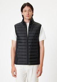 Gilet imbottito nero con chiusura a zip, colletto alto e due tasche laterali. Presenta cuciture orizzontali per l'isolamento. Indossato sopra una camicia bianca.