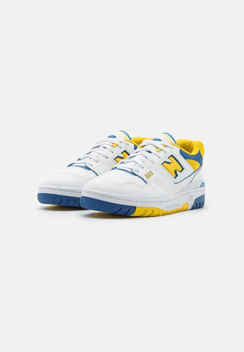 Weiße Sneakers mit blauen und gelben Akzenten, gelochtem Textil, runder Zehenkappe, Schnürsystem und einer dicken Gummisohle mit Farbblöcken.