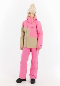 Rosa und beige Skijacke mit hohem Kragen und Reißverschlusstaschen, kombiniert mit passenden rosa Skihosen; schwarze Schneestiefel.