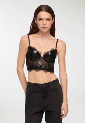 Bralette in pizzo nero con ricami floreali e bordi ondulati, dotato di spalline regolabili e design cropped, abbinato a jeans neri a vita alta.