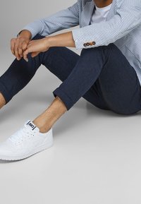 Bílé tenisky, tmavé nohavice s ohrnutými lemy a modro-bílé pruhované sako, sedící s rukama složenýma na neutrálním pozadí.