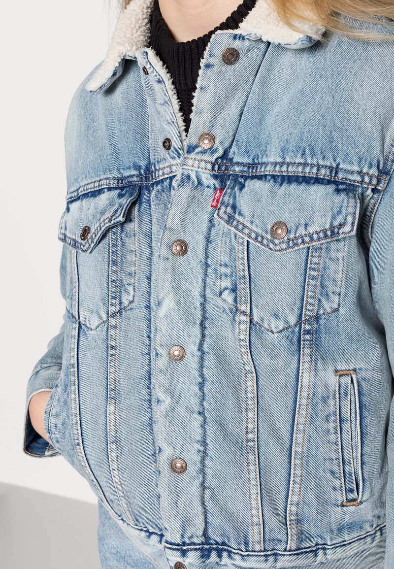 Veste en denim bleu clair avec un col en fourrure, boutons en argent, deux poches poitrine et détails de coutures. Texture douce avec une coupe décontractée.