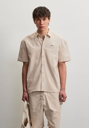 Jeune homme aux cheveux effet mouillé porte une chemise beige à manches courtes boutonnée et un short assorti, tenant un sac en tissu beige.