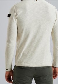 Pull beige à manches longues avec un motif en maille texturée, comprenant un détail de bouton à l'arrière du col et un petit accent noir sur la manche.