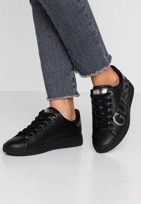 Zwarte synthetische sneakers met een gladde textuur, voorzien van logo-accenten en metalen details, gecombineerd met grijze jeans met een rafelige zoom.