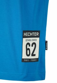 Blaues Baumwoll-T-Shirt mit einem schwarz-weißen Etikett, auf dem "HECHTER", "62" und "PARIS - FRANCE" gedruckt sind. Kurzärmlig mit rundem Ausschnitt.