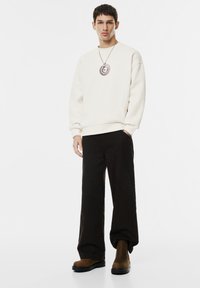 Mladý muž oblečený v bílém svetru, černých širokých kalhotách, hnědých botách a velkém kulatém pendantovém náhrdelníku, stojící na bílé pozadí.