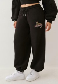 Persona valkā melnos sporta šortus ar pusdārgakmens leoparda un "True Religion" logo, baltas kurpes un īsu melnu topu.