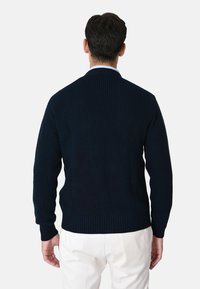 Maglione navy lavorato a maglia con texture a coste, caratterizzato da un collo alto e maniche lunghe, indossato sopra pantaloni bianchi. Vista posteriore che mette in evidenza il design senza cuciture.