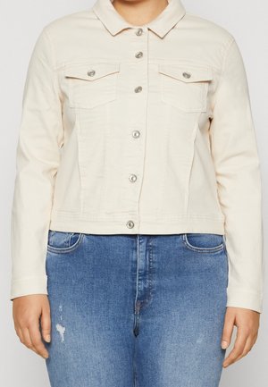 Frau trägt eine cremefarbene, mit Knöpfen versehene Jeansjacke mit Brustklappentaschen und blaue Jeans mit leichter Abnutzung.