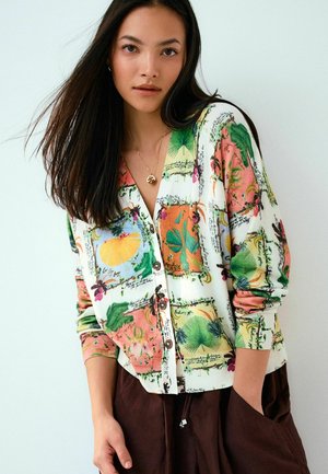 Žena s dlhými tmavými vlasmi v bielom kardigane na gombíky s farebnými potlačami listov a ovocia a v tmavohnedých nohaviciach pózuje s jednou rukou v vrecku.