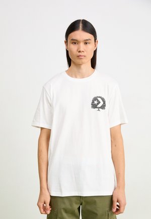 Camiseta de algodón blanca con un diseño gráfico negro que asemeja un árbol en el área izquierda del pecho. Cuello redondo y mangas cortas.