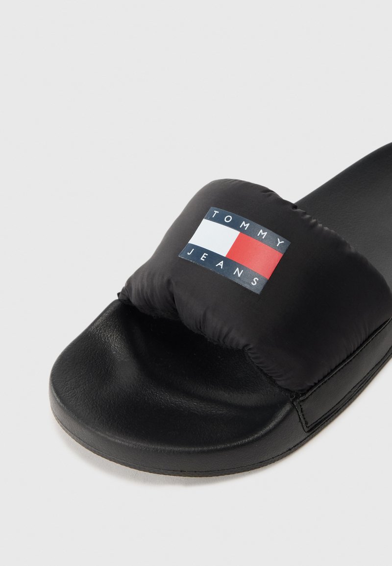 Schwarze Rutsche-Sandalen mit einem gepolsterten Riemen, der ein Logo mit roten, weißen und blauen Rechtecken zeigt. Hergestellt aus synthetischen Materialien, flacher Sohle.