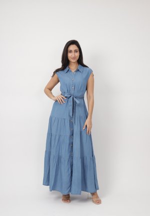 Kobieta ubrana w bezrękawową, zapinaną na guziki, jasnoniebieską długą sukienkę dżinsową z wiązanym w pasie paskiem, stojąca na tle gładkiej białej ściany.