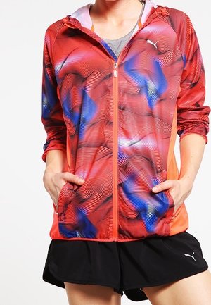 Vrouw draagt een rood-blauw-zwart patroon zip-up hoodie met handen in de zakken en zwarte Puma short.