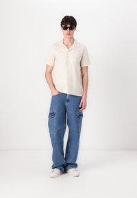Helles beigefarbenes Kurzarmhemd mit strukturiertem Muster, kombiniert mit übergroßen blauen Jeans-Cargohosen mit Seitentaschen und weißen sneakers.