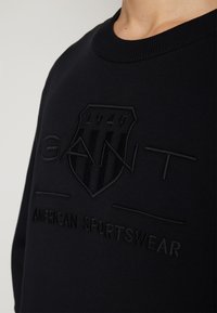 Sweat-shirt noir avec logo GANT en relief, emblème en forme de bouclier avec "1949" et texte "AMERICAN SPORTSWEAR" sur la poitrine.