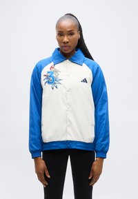 Γυναίκα με μακριά αλογοουρά που φοράει λευκό και μπλε μπουφάν με παπαγάλο, αριθμό 3 και λογότυπο Adidas, στέκεται μπροστά σε απλό φόντο.