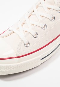 Gros plan d'une basket blanche en toile avec des lacets blancs, des œillets argentés, une bande rouge au-dessus de la semelle en caoutchouc et un bout renforcé blanc texturé.