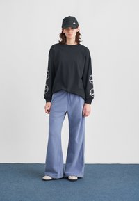 Mladý človek oblečený v čiernom tričku s dlhými rukávmi s bielymi vzormi na rukávoch, modrých širokých nohaviciach, čiernej šiltovke Nike a bielych teniskách stojí na modrom koberci.