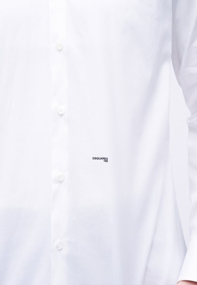 Chemise blanche à boutons avec un petit logo noir "DSQUARED2 1995" sur le côté gauche près des boutons, manches longues visibles.