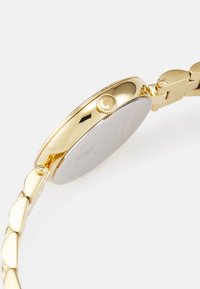 Gold-tone wristwatch with a round case, smooth bezel, and linked bracelet. Features a prominent crown and a reflective silver back.